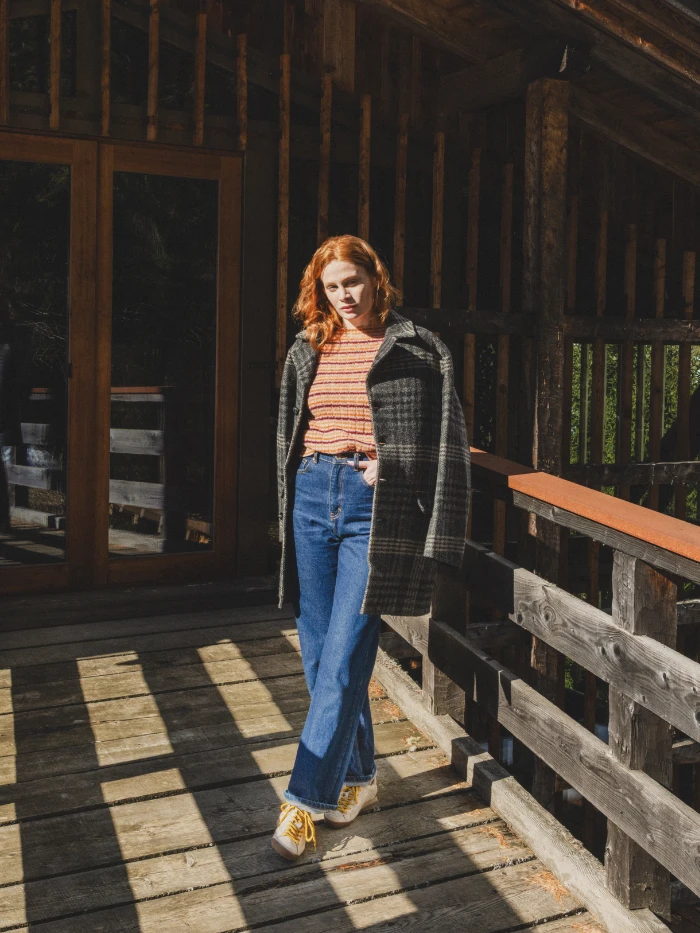 Woman wearing a plaid coat, striped top, and wide-leg jeans posing on a wooden mountain balcony