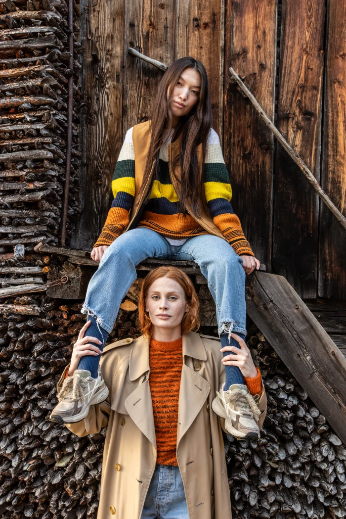 Two models wearing autumn outfits with striped sweaters, trench coats, and sneakers in front of a wooden woodpile